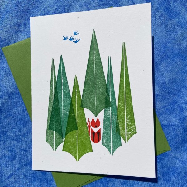 An image of a letterpress printed card. The card is printed with simple geometric shapes in a design featuring a group of pine trees in different shades of green, and a red fox sitting among the trees.