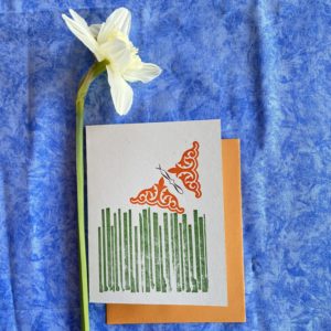 a light blue card with an orange envelope on a blue background. On the card is printed in a geometric style grass of various lengths, and flying over the grass an orange butterfly composed of typographic ornaments. next to the card is a daffodil.