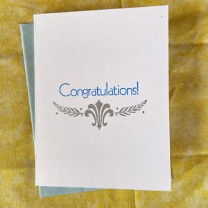 a white card on a yellow background. The card reads “congratulations!” in blue letters. Underneath this are silver laurel branches and a stylized fleuron.